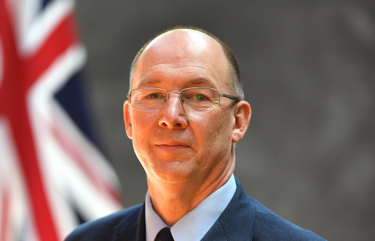 Wing Commander Graham Streatfield in full military dress uniform, standing in front of the New Zealand flag smiling.