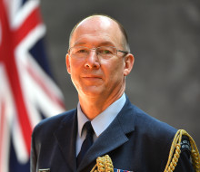 Wing Commander Graham Streatfield in full military dress uniform, standing in front of the New Zealand flag smiling.