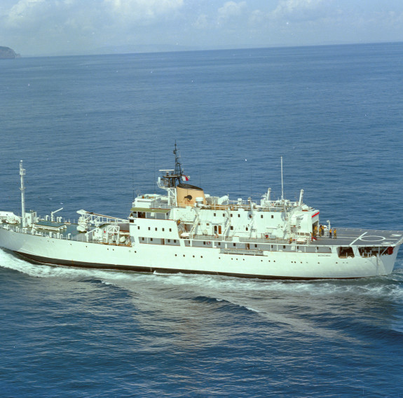 A vintage photos of the actual HMNZS Monowai at sea in 1979. The ship is travelling to the left of the frame with land and the horizon visible in the background.