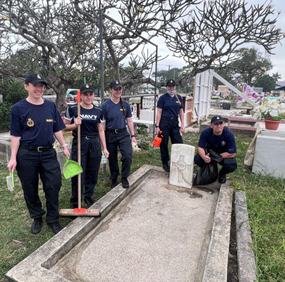 Five people in Navy blue t-shirts, caps and trousers clean a burial plot in Tonga. With brooms and brushes in hand, they stand in a cemetery surrounded by flowers and a large tree.