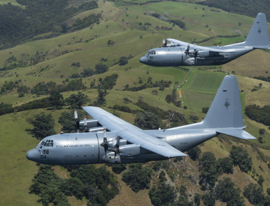 Two C-130H aircraft flying in close formation over hilly and sparsely wooded farmland. They are seen from alongside, slightly ahead and above.