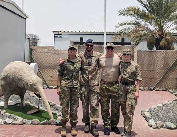 Four personnel, in camouflage military uniform have their arms around one another as they smile at the camera in front of a New Zealand flag and a large sculpture of a kiwi.