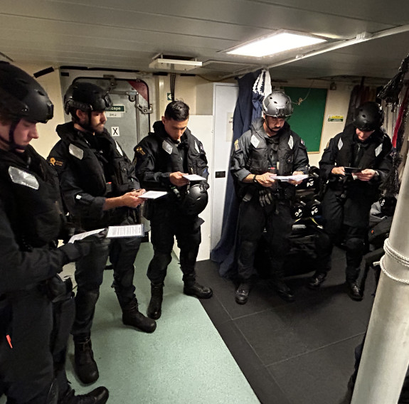 The Deployable Boarding Team brief aboard HMS Spey prior to the boarding exercise. Each of the five sailors sailors in uniform are reading from paper.