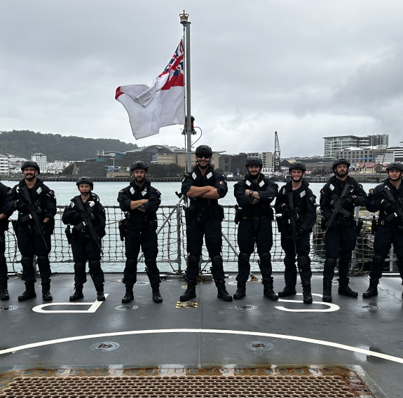 The Deployable Boarding Team stand on the flight deck of HMS Spey with their weapons. In the background is Wellington city, where the team disembarked the ship/