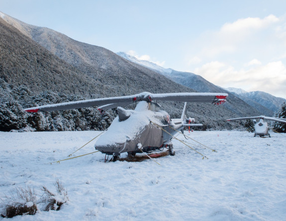 Two A109 helicopters with covers and tie-down ropes attached, are covered in snow at Dip Flat in winter.