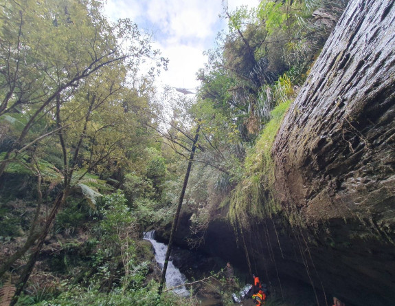 An NH90 helicopter hovers over the area where they are about to pick up a patient. There is a lot bush and a waterfall. 