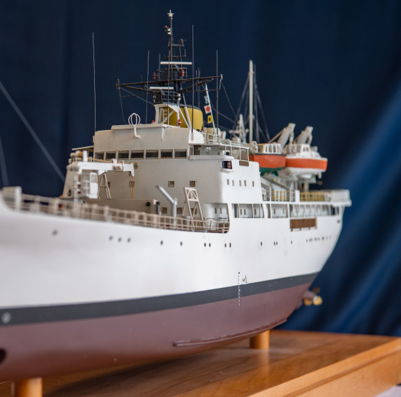 A front-on view of the model of HMNZS Monowai which sits on a wooden stand. The ship is white with a black Plimsoll line and below that is brown. The model has intricate details including portholes, masts and cables.