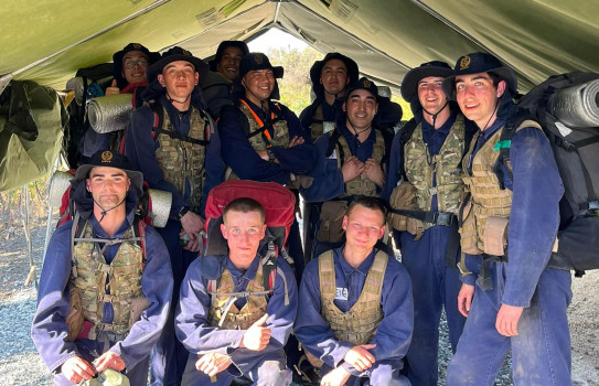 Sailors in training smile for a photo in a tent, all wearing blue overalls, camouflage vests and tramping pack.