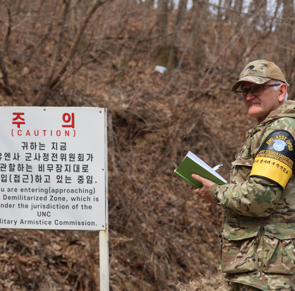 A soldiers stands next to a sign in front of brown dried up trees and branches.
