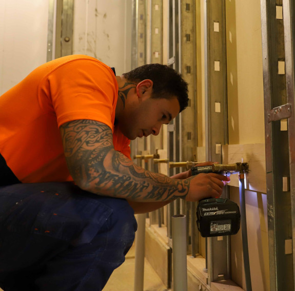 LCPL Spencer uses a drill to secure a hose to the back of a wall. He is wearing an orange t-shirt and navy blue work pants. He also has tattoos on his right arm.