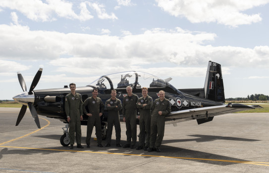 Six Black Falcon pilots stand in front of the black Texan aircraft. They are wearing green overalls as they stand on the tarmac at Base Ohakea with scattered cloud and blue sky behind them.