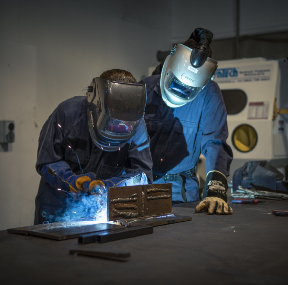 A teacher under supervision gets hand-on welding experience. Both the teacher and supervisor are wearing protective overalls and masks as sparks fly from the welding.