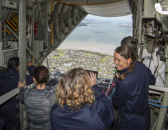 The view from on board a C-130H (NZ) during flight. Four teachers are in the foreground with the rear door open and the Hauraki Gulf in the background.