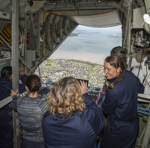 The view from on board a C-130H (NZ) during flight. Four teachers are in the foreground with the rear door open and the Hauraki Gulf in the background.