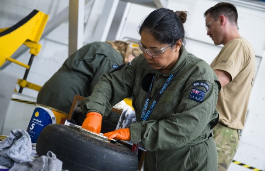 A female wears a green military flying suit and orange disposable gloves as she works on a tyre.  The woman has dark hair tied in a bun, is wearing a lanyard and safety glasses while she works.  In the background there is a male dressed in military attire