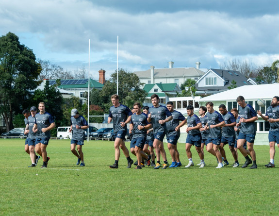 Te Taua Moana rugby team training on a green field. The players are all running in the same direction, in uniform. 