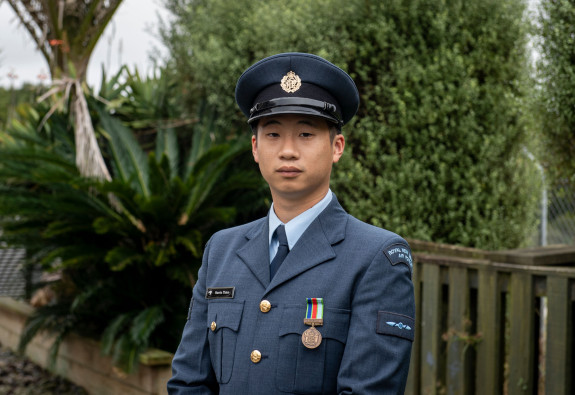 Portrait image of a man named Harris wearing formal blue Air Force uniform with a hat standing outside near a fence and some green bush. 