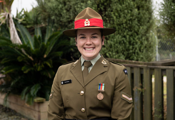Portrait of a female army soldier wearing formal Army uniform with a headress. She's smiling and is standing outside in front of some greenery. 