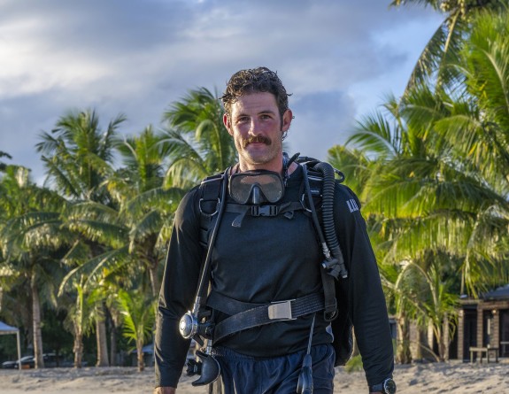 A diver walks along a beach while wearing full diving equipment and holding fins. In the background, white sand, meets the water and trees follow the coastline.