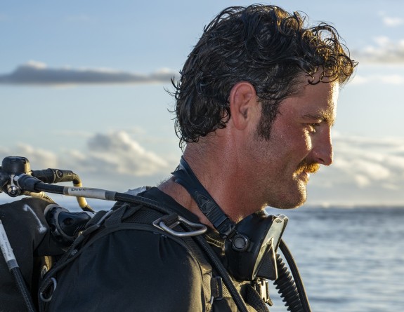 Lieutenant Christian Lloyd in front of the ocean, blue skies with fluffy clouds on the horizon. He is equipped to go diving.