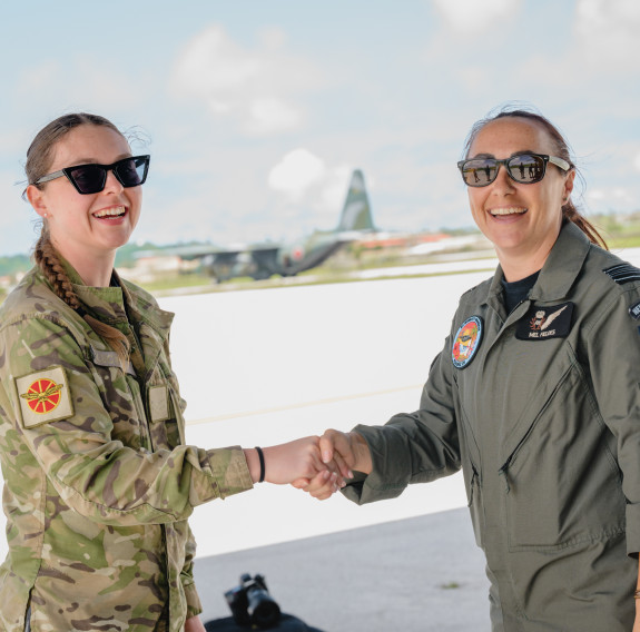 Two women in military uniforms shake hands on the tarmac with an aircraft in the background.