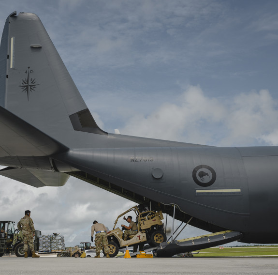 A military vehicle drives out the back of a large grey aircraft that has a kiwi roundel on the side.