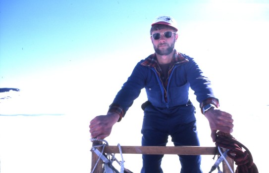 A man holds onto the handles of a sled, he wears ski type clothing, sunglasses and a cap with the blue sky and snow behind him.