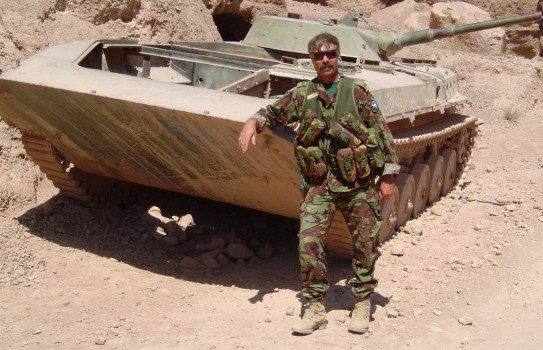 A soldier poses alongside a military tank like vehicle in desert surroundings.