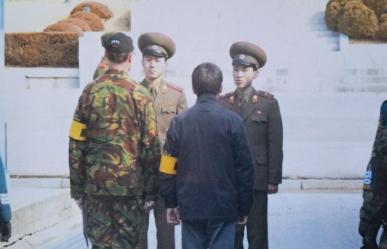 Two Korean soldiers stand in dress uniform, hats atop directly opposite a New Zealand army soldier, black cap on and yellow armband over a military camouflaged uniform. Beside him he is flanked by a shorter male in civilian clothing.  They are standing at