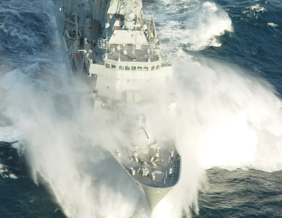 HMNZS Te Kaha soars through a large wave, sea spray encompasses the bow of the ship, reaching the bridge.