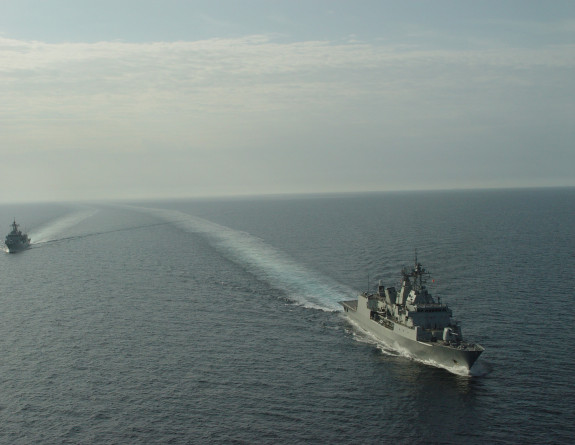 HMNZS Te Kaha sailing over smooth seas, the wake of the ship stretches to the horizon.