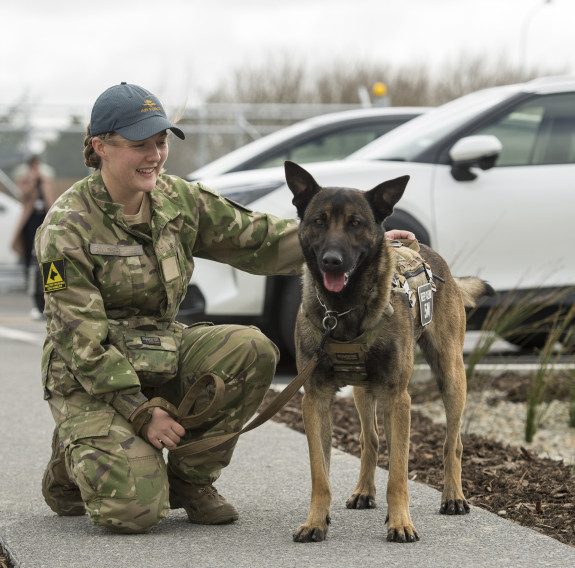 An aircraftman dog handler knells with their dog. The dog is looking directly at the camera and the handler is smiling, looking at the dog.