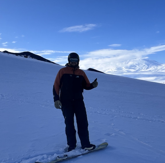 CPL Jobling, left food attached to a snowboard and a thumbs up, stands in the shadow of a mountain on the ski slopes. Plenty of snow covers the side of the mountain with a couple of rocks appearing from the snow near the edge. In the background it's much 