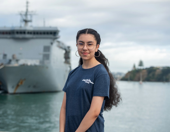 Maria La Parra, a School to Seas participant stands in front of HMNZS Canterbury, a large grey ship at Devonport Naval Base, she is wearing a blue 'School to Seas' branded t-shirt and large hooped earrings.