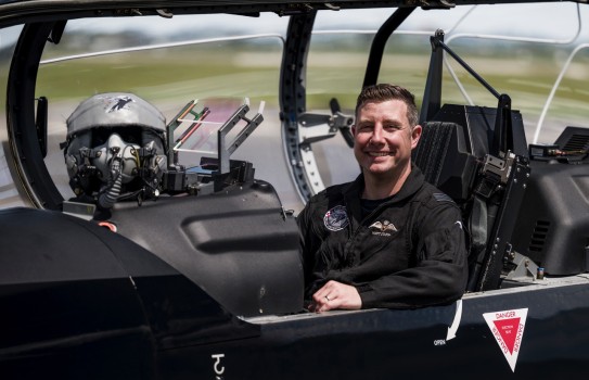 A male wearing a black flying suit sits inside the cockpit of a small aircraft with the hood open. His hand rests on the side of the aircraft whilst looking at the camera.