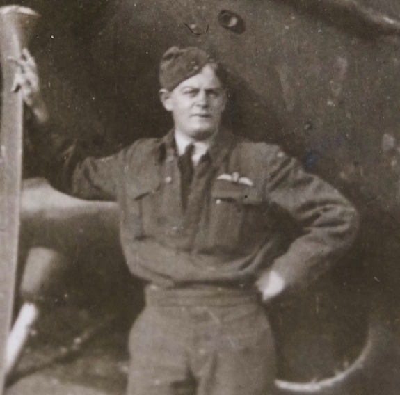 A sepia photo of a man in Air Force uniform with one had on the propellor of a Spitfire and his other hand on his hip.