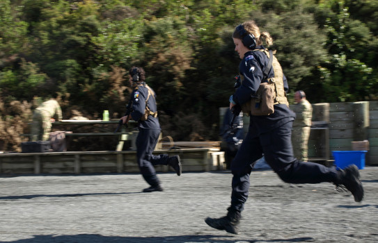 Two NZDF personnel moving swiftly at the shooting range with their weapons. Both wearing dark blue overalls, ear protection and an ammunition vest.