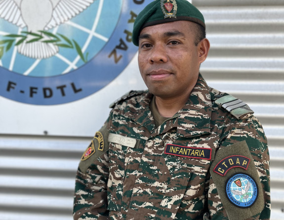 A solider stands in uniform - looking at the camera and smiling. In the back ground is corrugated iron and a sign. 