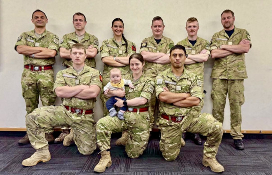 A group of soldiers in uniform look at the camera, in group formation. All have their arms crossed - the middle soldier in the front row (out of two rows) is holding a baby. 