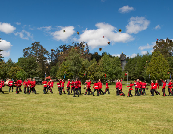 Soldiers in red and black dress uniform through their hats in the air as part of their graduation ceremony. They are standing on the grass on a sunny day. There is blue sky and very few clouds. In the background, there are trees.