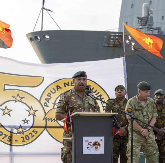 Papua New Guinea soldiers stand in front of a ship and the countries flag as one soldier stands at a podium, talking into a microphone.