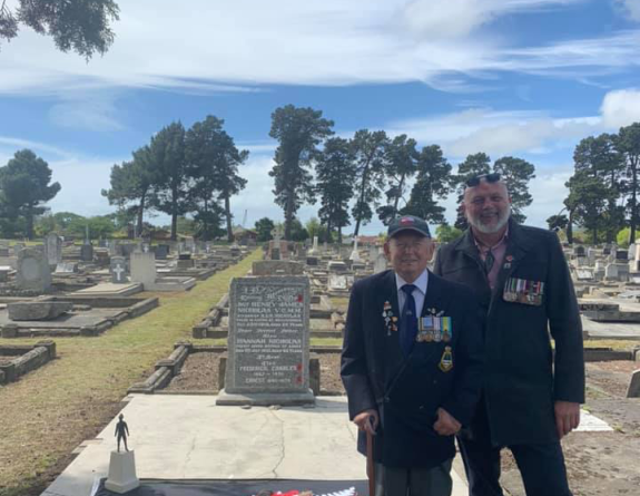 Two people stand looking at the camera smiling. One with a walking stick. In the background there are headstones and the rest of the cemetery. Next to them is a fern flag lying on the grave sit they are standing in front of.