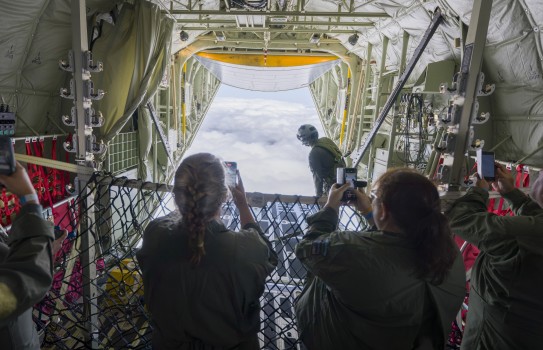 Two females wearing flying suit stand at the back of an aircraft looking out to the cloud beyond the open rear. They stand holding their phones taking photos of the cloudy scene beyond, a cargo net acts as a barrier between them and the opening and a mili