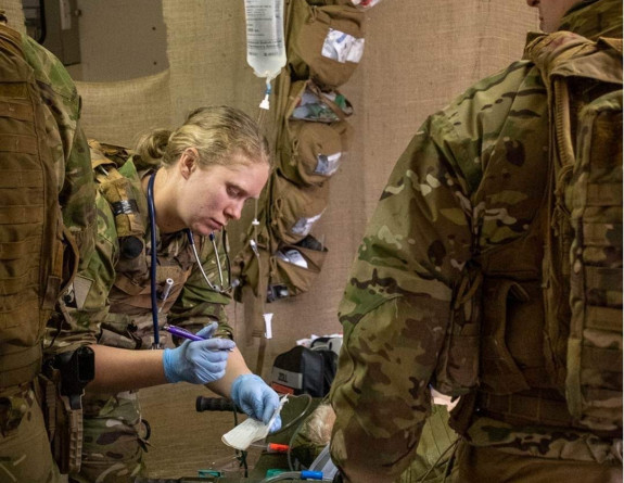 A nurse write notes with paper and a pen in an operating environment while working with a patient. They are in a tent and two other personnel, also wearing camouflage uniform stand are moving in the forground.