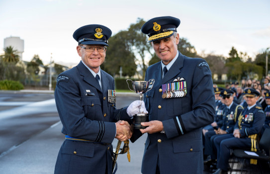 Two aviators shake hands, while exchanging a trophy while smiling at the camera.