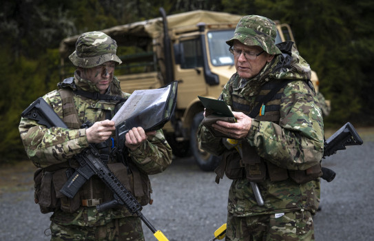 Two aviators in full camouflage uniform read paperwork and write in a notebook with weapons slung around them. In the background is a large tan military truck.