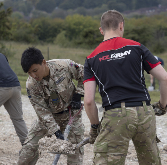 Four people in Army uniform, one of which is wearing a red and black New Zealand Army shirt, help distribute chalk. Green grass, bushes and trees fill the background.