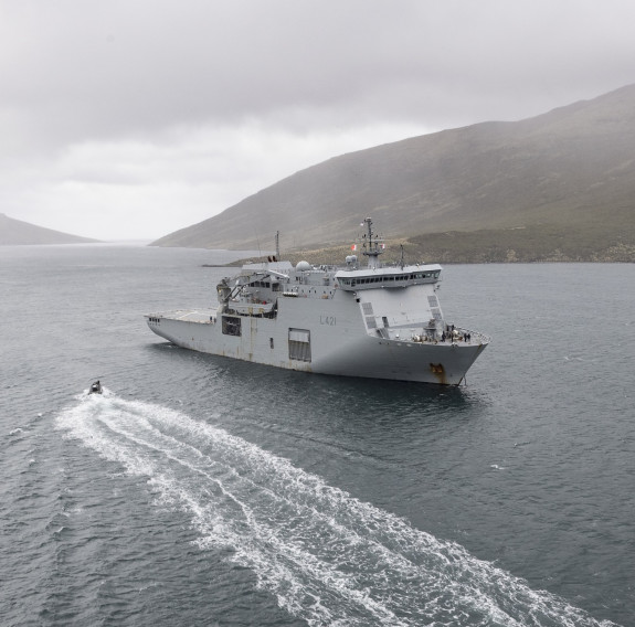 HMNZS Canterbury amongst the islands as a RHIB creates a large wake as it travels towards the ship. The clouds are low and rain falls on the brown rolling hills in the background.