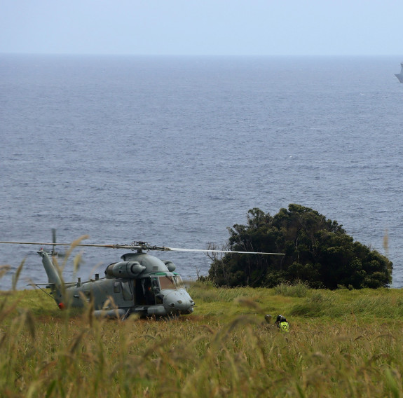 A grey Seasprite helicopter sits on the land with the ocean spanning to the horizon. HMNZS Canterbury waits at sea, just below the horizon on an overcast day.