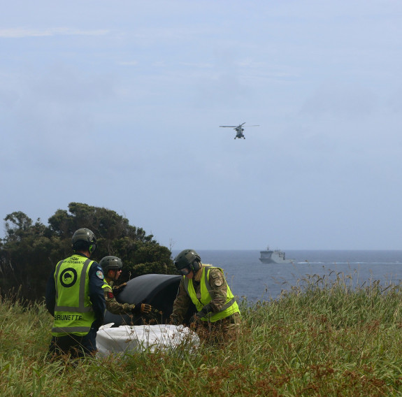 Personnel in high vis vests and uniform work on land as a Seasprite helicopter approaches over HMNZS Canterbury.